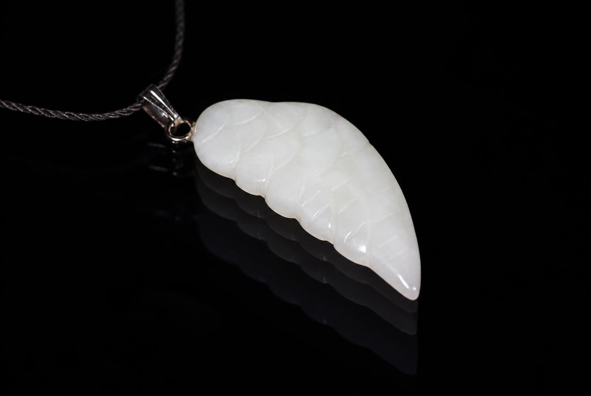 A white jade pendant shaped like a wing with carved lines, hanging from a silver-toned bail on a dark cord against a black reflective surface.