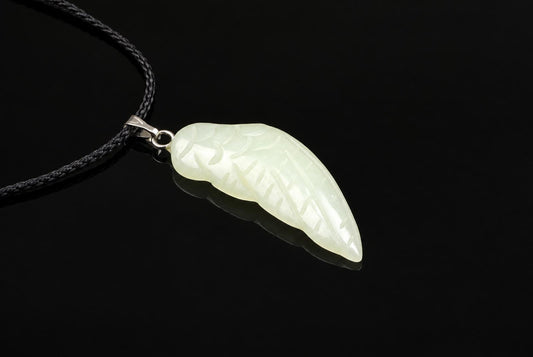 A pale green jade pendant carved into a wing shape with engraved lines, attached to a black cord via a silver-toned bail on a dark surface.