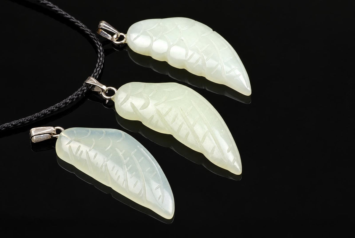 Pale green jade wing pendants show carved feather patterns, arranged with silver toned loops and a black cord on a dark surface.