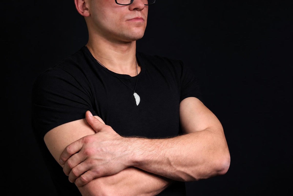 A man in a black t-shirt wears a white, wing-shaped pendant on a dark cord against a black background.