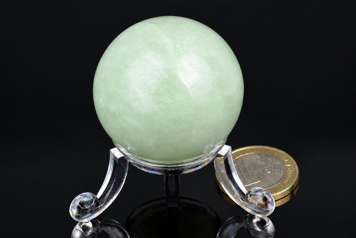 A pale green, smooth sphere rests on a clear acrylic stand against a dark surface.