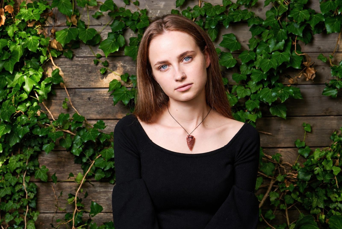 A woman wears a dark red arrow-shaped pendant on a thin cord against a wooden wall with green ivy.