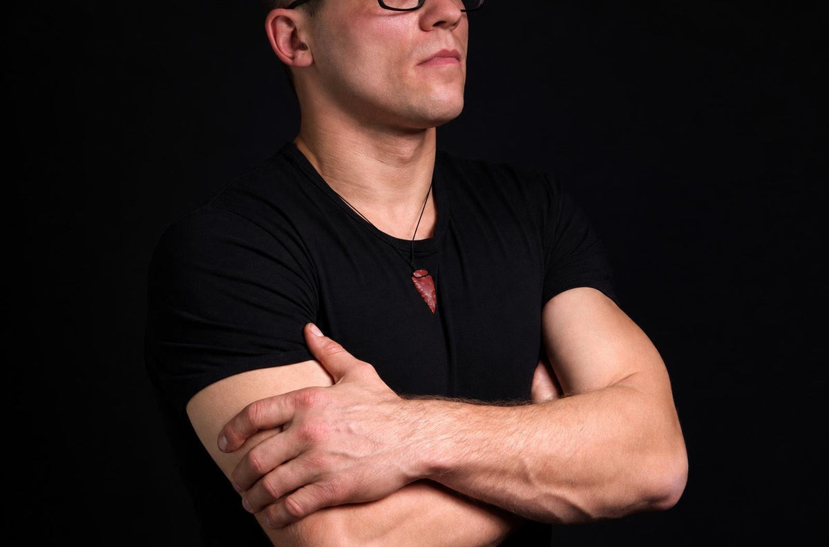 A man in a black t-shirt wears a dark red arrow-shaped pendant on a thin cord against a dark background.