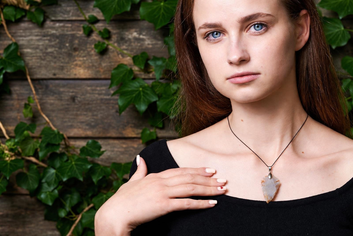 A woman wears a necklace with an arrow-shaped jasper pendant on a black cord, against a wooden background with green ivy.