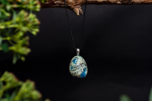 A teardrop-shaped pendant with a silver-toned bail hangs from a black cord against a dark background, featuring blue and white speckled stone.