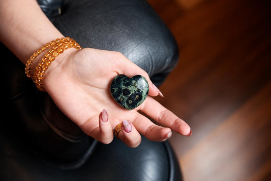 A hand holds a polished, heart-shaped stone with dark green and black swirling patterns on a dark leather surface.