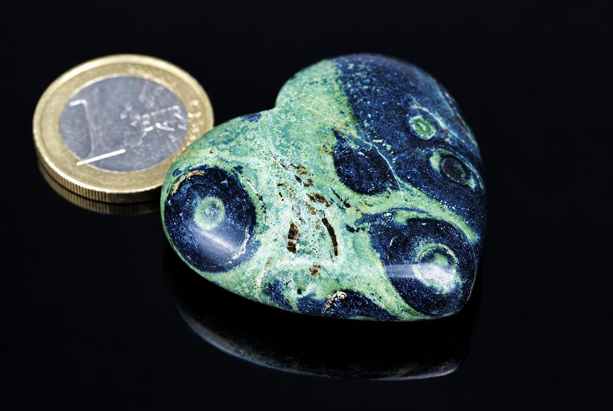 A polished green heart-shaped stone with dark circular markings rests beside a coin on a reflective black surface.