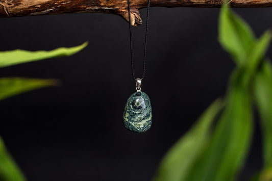 A polished green pendant with light speckles hangs from a black cord on a wooden branch, framed by blurred green leaves.