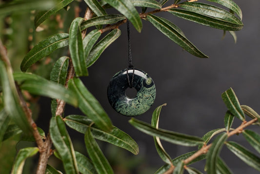 A polished, dark green and black patterned donut-shaped pendant hangs from a black cord on a leafy branch.