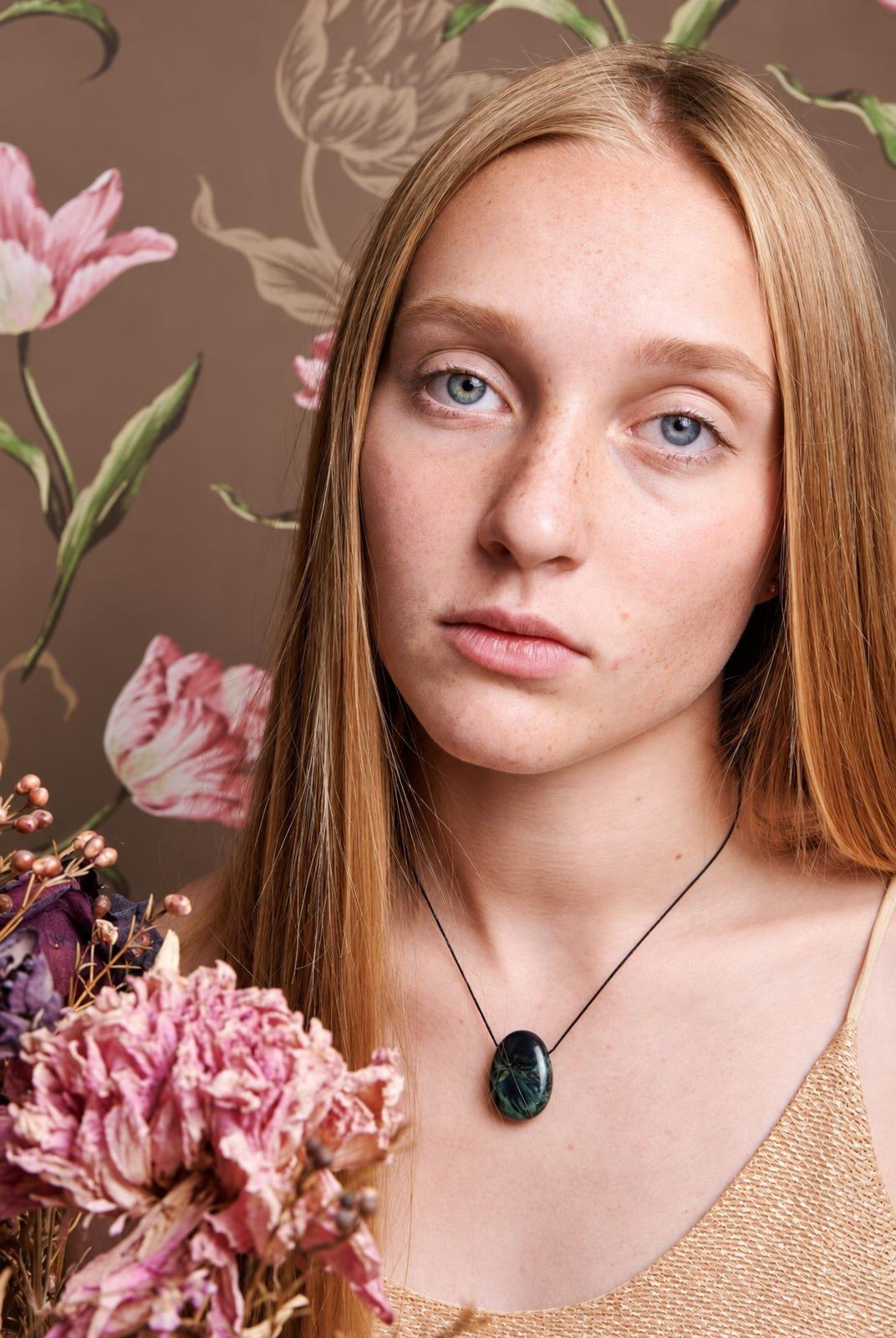 A woman wears a black cord necklace with an oval, dark green pendant featuring lighter veins against a floral background.