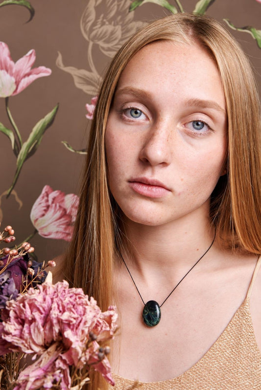 A woman wears a black cord necklace with an oval, dark green pendant featuring lighter veins against a floral background.