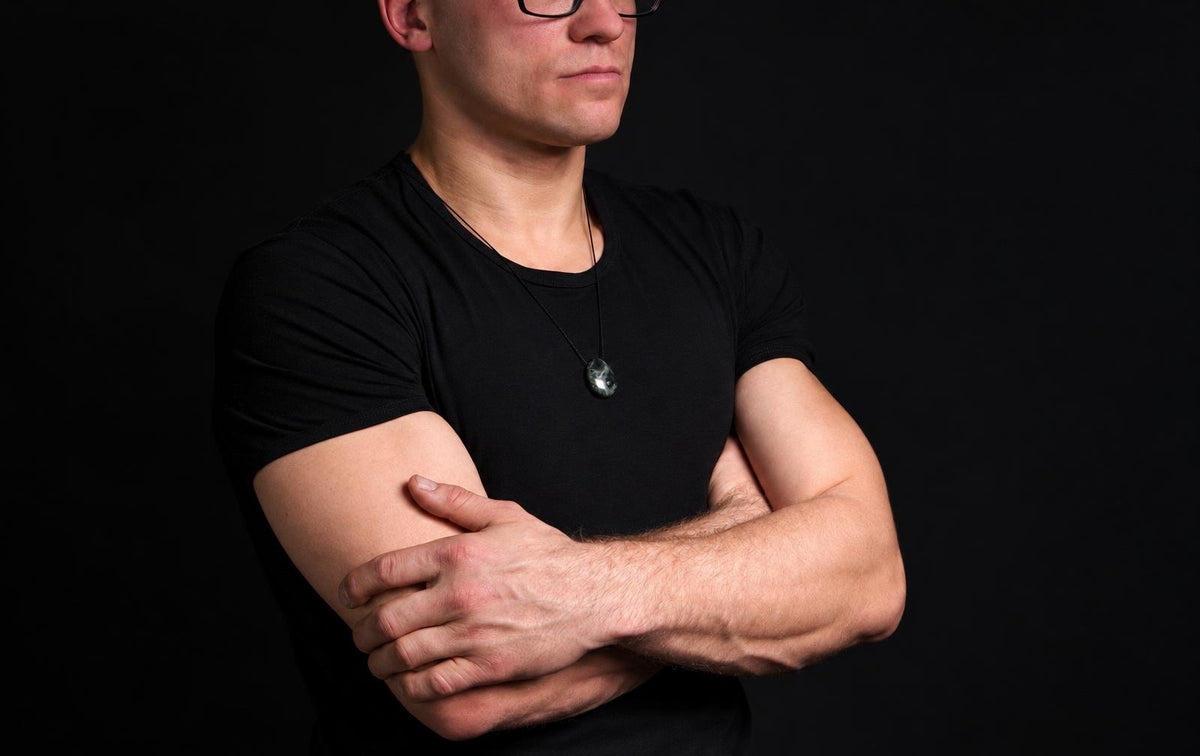 A man in a black t-shirt wears a dark green, oval-shaped pendant on a thin cord against a dark background.