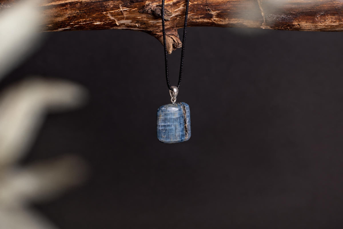 A blue kyanite pendant with a silver bail hangs from a black cord on a dark wooden branch against a dark background.