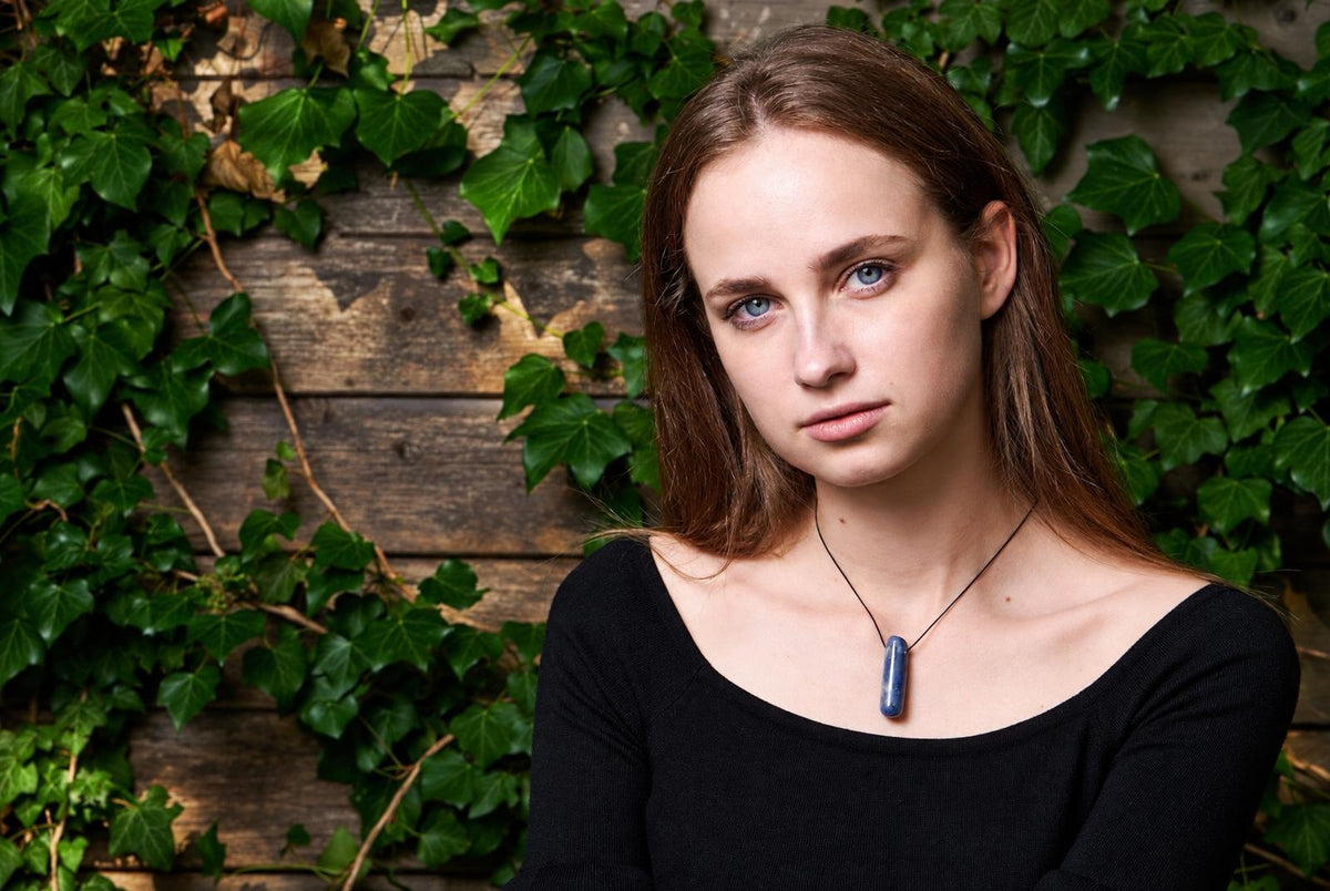 A woman wears a dark blue, elongated pendant on a thin cord against a wooden wall with green ivy.