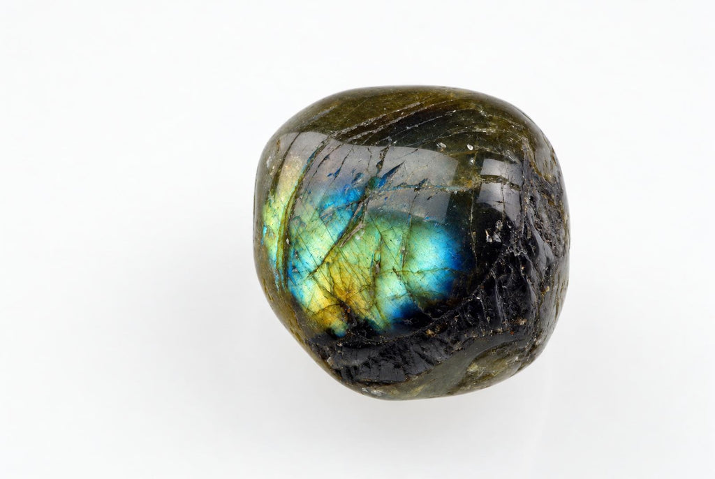 A rounded polished dark grey stone with blue, green and yellow flashes and fine dark lines rests on a white background.