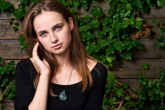 A woman wears a black top and a pendant with a spiral ammonite shape in greenish-blue labradorite against a wooden wall with ivy.