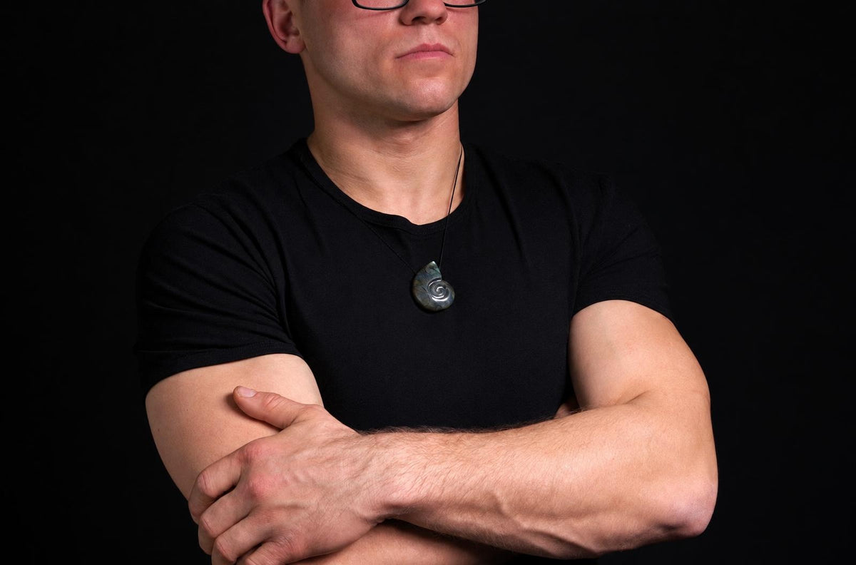 A man wears a dark t-shirt and a pendant with a spiral ammonite shape in grey-blue tones against a black background.