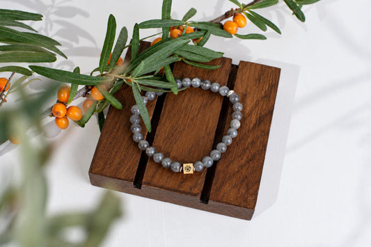 A bracelet of gray round beads with a small square metal bead rests on wooden slats beneath a branch with narrow leaves and orange berries.