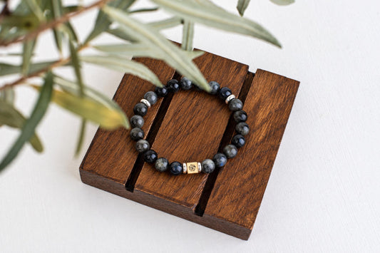 A bracelet of dark gray round beads with a small square metal bead rests on wooden slats beneath green leaves.
