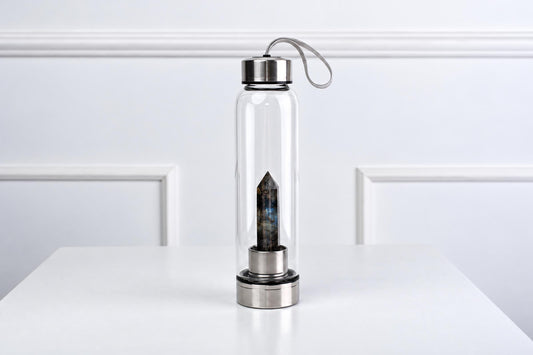 A clear glass water bottle with a silver-toned lid and base, containing a dark blue-grey obelisk crystal on a white surface.