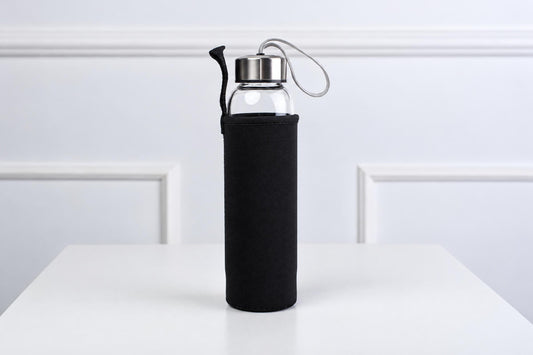 A clear glass water bottle with a silver-toned lid and black fabric sleeve, standing on a white surface.