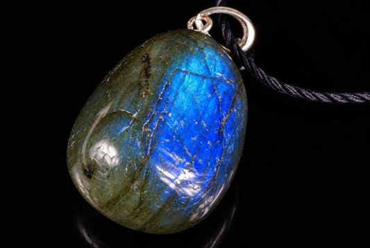A polished Labradorite pendant with blue and green iridescence, set in a silver-toned bail on a black cord against a dark background.