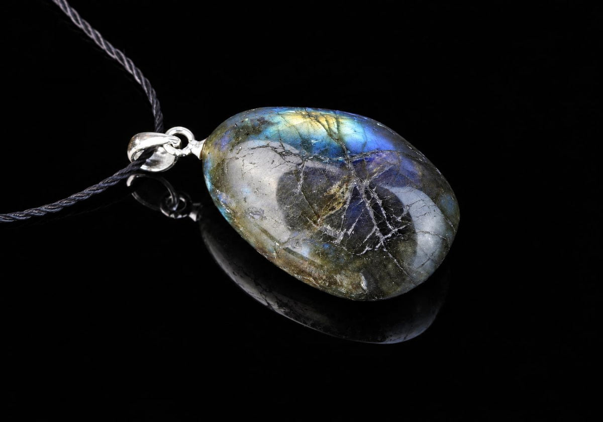 An oval labradorite pendant shows blue and yellow flashes across a dark mottled surface, framed by silver-colored metal against a black background.