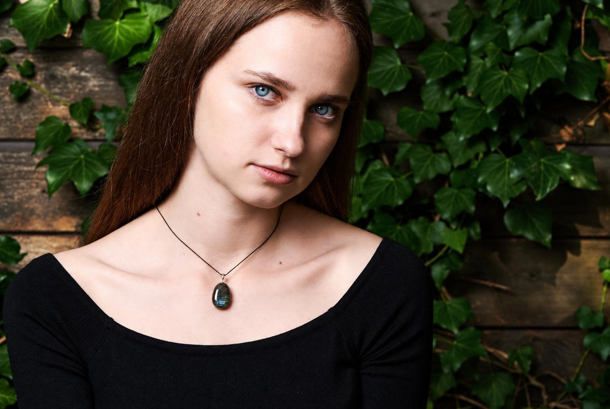 A woman wears a black top and a pendant with a dark, blue-green stone on a thin cord against a wooden background with ivy.