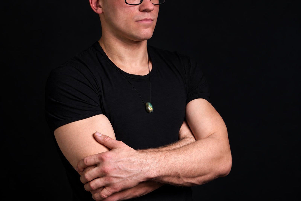 A man in a black t-shirt wears a Labradorite pendant on a dark surface against a black background.