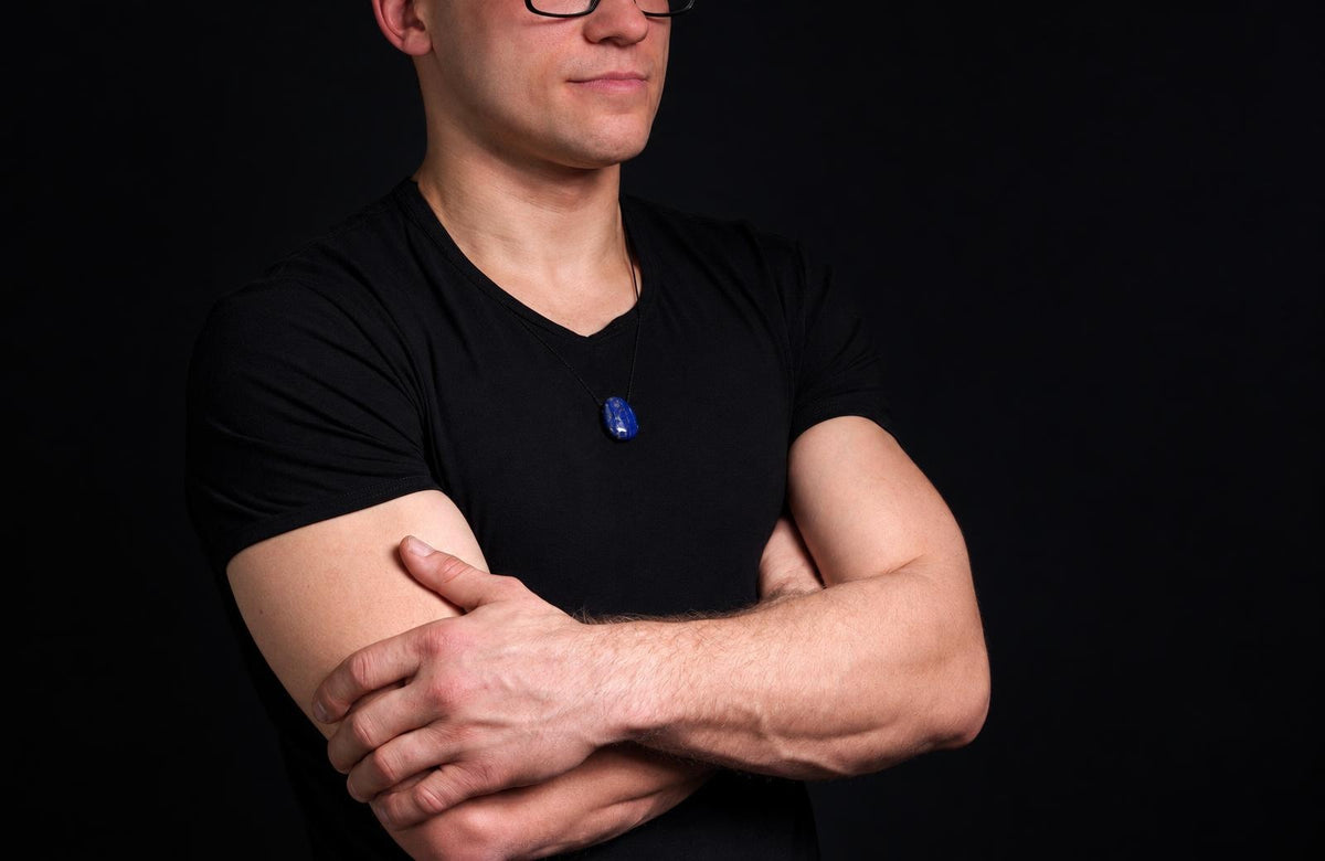 A man in a black t-shirt wears a necklace with a dark blue, oval pendant against a dark background.