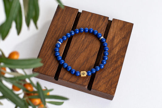 A bracelet of blue round beads with a small square metal bead rests on wooden slats beside green foliage on a white surface.