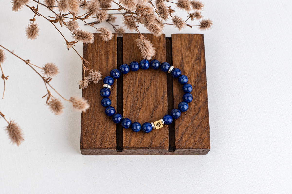 A blue beaded bracelet with silver and gold-toned accents rests on a dark wooden surface, surrounded by dried flowers.