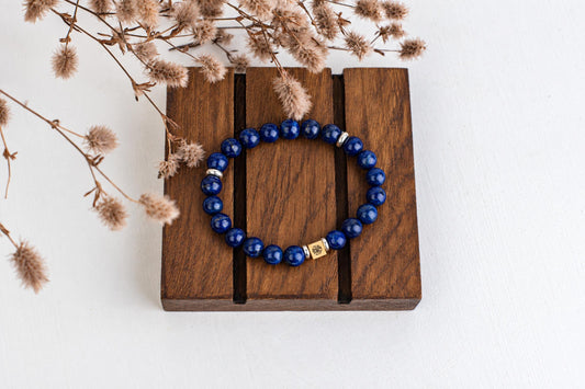 A blue beaded bracelet with silver and gold-toned accents rests on a dark wooden surface, surrounded by dried flowers.