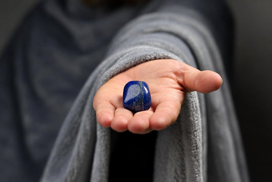A person in a grey robe holds a polished, deep blue stone with golden flecks in their open palm against a dark background.