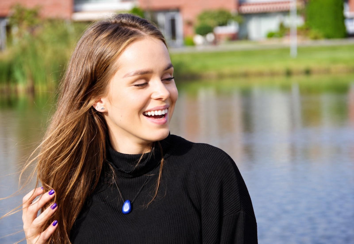 A smiling woman with long brown hair wears a black turtleneck and a blue teardrop pendant necklace against a blurred water background.