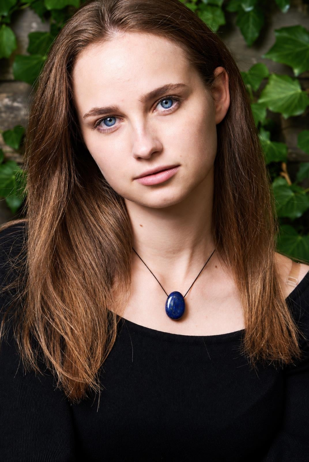 A woman wears a black top and a dark blue oval pendant on a thin cord against a green leafy background.