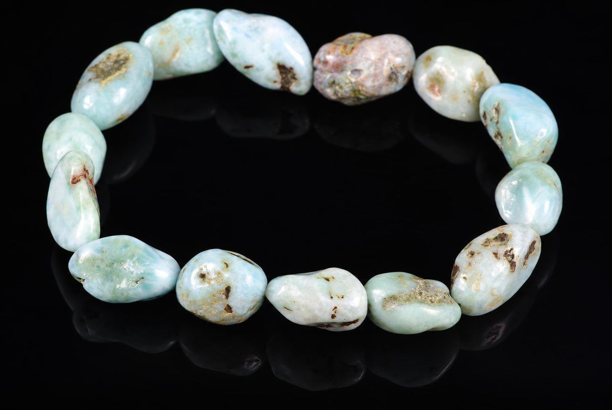 A Larimar bracelet with smooth, rounded beads in pale blue and white tones, some with brown or pinkish spots, arranged on a dark reflective surface.