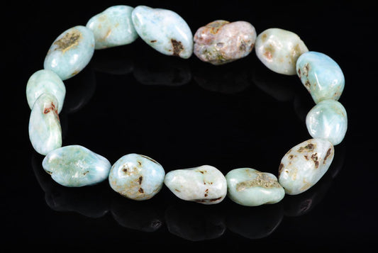 A Larimar bracelet with smooth, rounded beads in pale blue and white tones, some with brown or pinkish spots, arranged on a dark reflective surface.