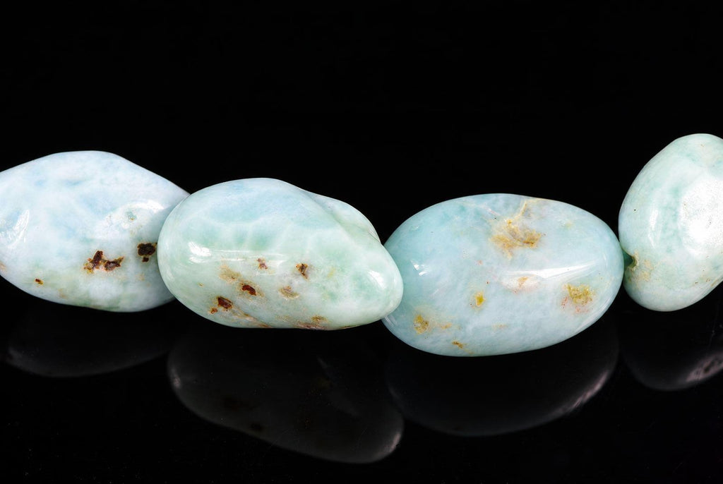 Four polished Larimar beads in pale blue-green with brown and yellow spots, arranged on a dark reflective surface.