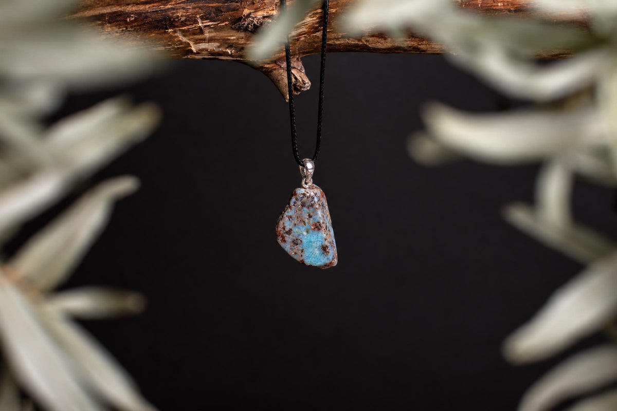 A Larimar pendant with a blue and brown stone hangs from a black cord on a wooden branch against a dark background.