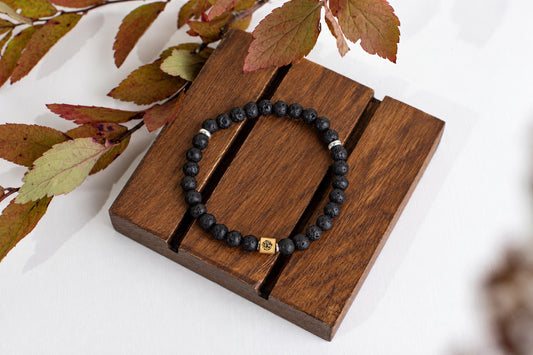 A black lava stone beaded bracelet with silver-toned spacers and a gold-coloured square charm rests on a dark wooden surface.