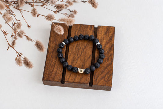 A black lava stone bracelet with silver-toned beads and a gold-coloured square charm rests on a dark wooden surface.