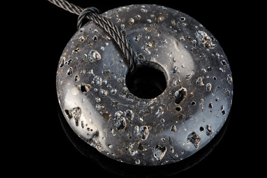 A dark, donut-shaped pendant with a central hole and a braided cord. Its surface is speckled with lighter inclusions on a black background.