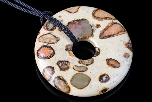 A donut-shaped pendant with a cream base and embedded brown, orange, and grey mineral inclusions on a dark surface.