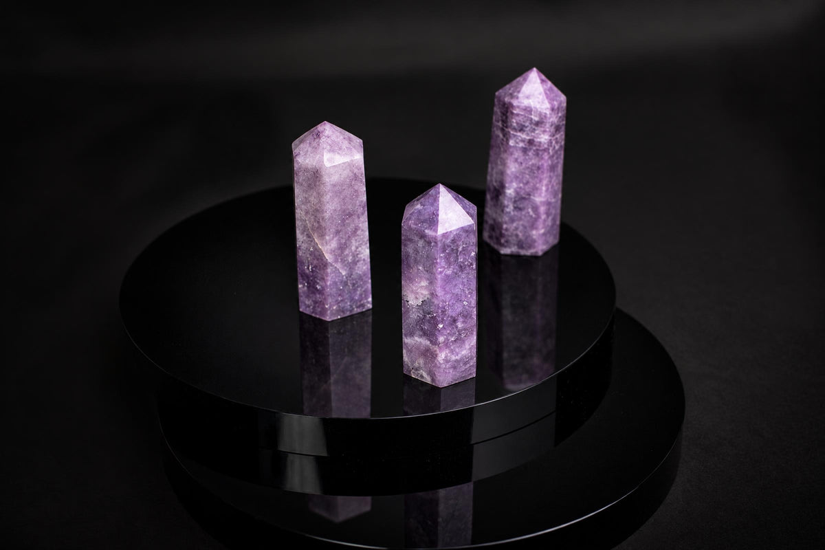 Three purple lepidolite crystal points with pointed tops stand on a dark, reflective circular surface.