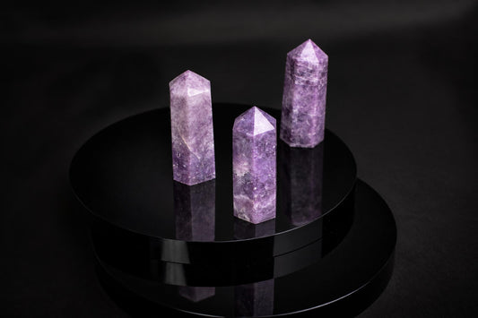 Three purple lepidolite crystal points with pointed tops stand on a dark, reflective circular surface.