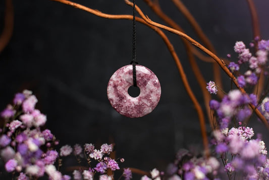 A pink and white speckled donut-shaped pendant hangs from a black cord against a dark background with purple flowers.