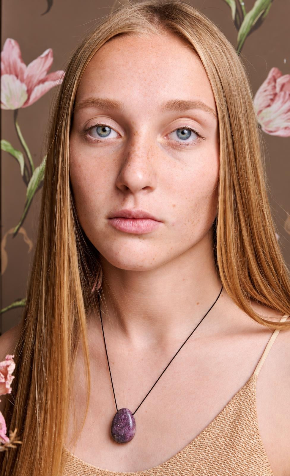 A woman wears a black cord necklace with an oval, dark purple lepidolite pendant against a floral background.