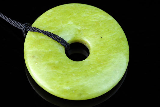 A round, pale green pendant with a central hole and a dark cord looped through it, set against a black surface.