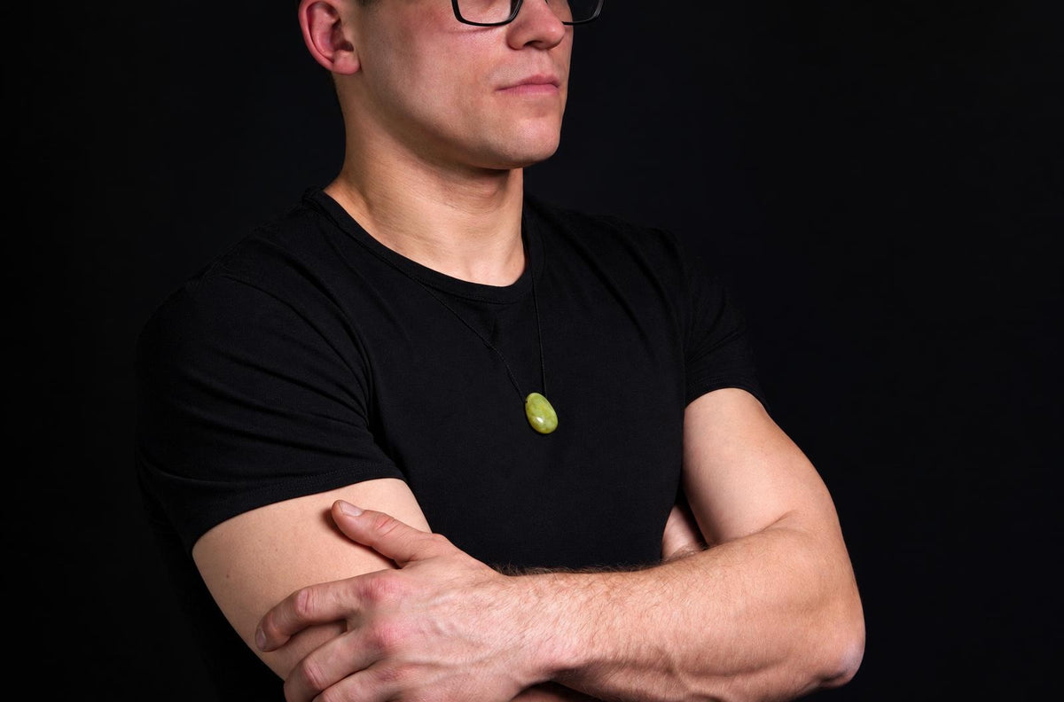 A man in a black t-shirt wears a necklace with an oval, light green pendant against a dark background.